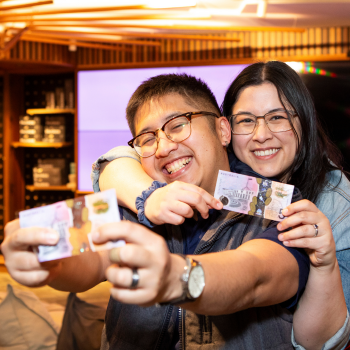 Two smiling people hold up Australian five-dollar notes while celebrating in a lively indoor setting with warm lighting and a modern background.